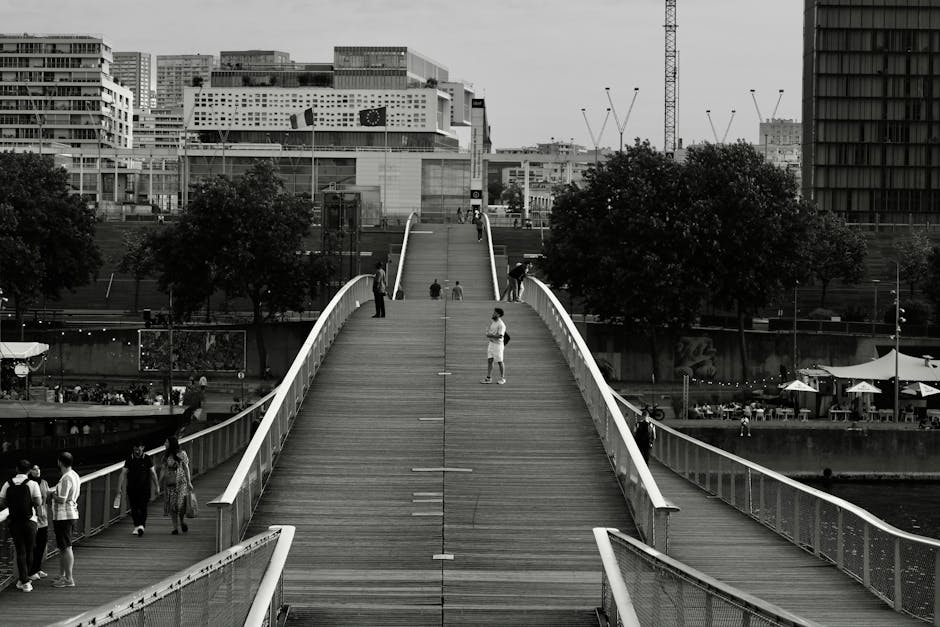 This image depicts a black and white view of a modern pedestrian bridge with a gently arched design, featuring white railings along both sides and a textured wooden walkway. Several people are walking