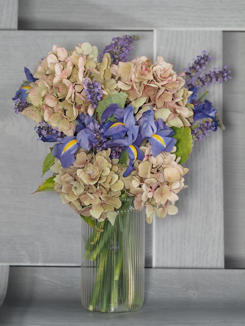 A tall, clear glass vase filled with a fresh floral bouquet featuring large, soft pink and cream hydrangea blooms, complemented by vibrant blue irises with yellow accents and sprigs of purple lavender