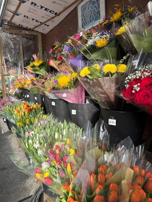 A display of various fresh flower bouquets at a flower shop, arranged in black buckets and wrapped in colorful paper. The arrangements include yellow chrysanthemums, purple and white daisies, red rose