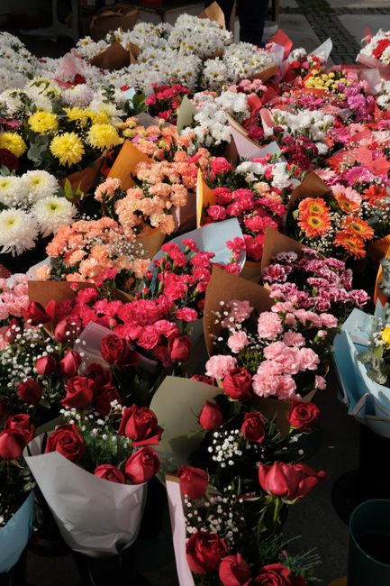 A vibrant display of fresh flower bouquets arranged closely together on a market stall, featuring a variety of blooms including red roses, pink carnations, white chrysanthemums, yellow daisies, and ba